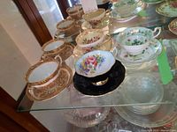 Photo showing arrangement of 5 teacups with matching saucers, featuring floral and gold patterns including one black saucer with floral teacup.