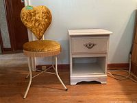 Photo shows vintage gold velvet upholstered chair with heart-shaped backrest next to white painted nightstand with drawer and open shelf on hardwood floor.