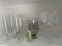 Photo showcasing 6 tall champagne flutes, 3 large balloon wine glasses, 6 small clear glass bowls stacked, and 3 small printed shot glasses on a shelf.
