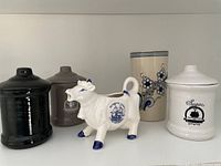 Photo showing white ceramic cow creamer and four assorted ceramic serving pots in black, gray-brown, cream with blue grapevine design, and white with sugar label and black emblem.