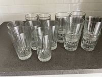Ten clear glass tumblers on a countertop, showing two sizes with vertical ribbed and textured designs, arranged in an upright position.