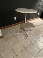 Side angle view of the round white laminate top bistro table and its metal base with four legs and adjustable feet on a tiled floor.