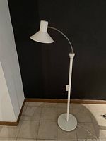 Full view of white MCM gooseneck reading lamp with round base and cone-shaped shade against a dark wall.