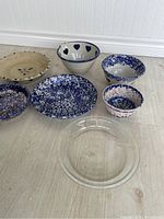 Photo showing six assorted pottery pie plates and bowls arranged on floor, including blue and white speckled ceramics, beige plate with floral motif, and clear glass pie plate