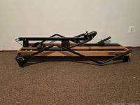 Side angled view of a wooden base ski exercise machine with black metal frame and foot straps, showing overall design and structure.