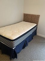 Twin bed setup showing woven headboard, mattress, and box spring with bed skirt in a carpeted room against a white wall.