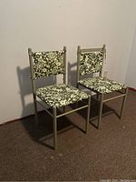 Two metal frame dining chairs with green floral patterned vinyl seats and backs, placed against a white wall on carpet.