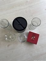 Photo of assorted glass vases, black plastic ice bucket, and small red box with wine stoppers on light wood floor