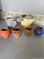 Photo showing collection of two watering cans and five plant pots on concrete floor against wall.