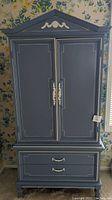 Front view of the blue Dixie armoire showing closed double doors with decorative white trim and handles beneath triangular pediment.