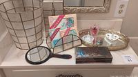 Full view of bath items arranged on bathroom vanity including jewelry box, mirror, perfume bottle, pill box, rice paper hankies, and decorative tray