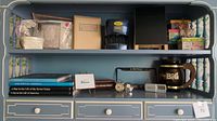 Wide view of shelf displaying books, clocks, black mini chalkboard, digital coin sorter, music box, decorative gift boxes, needlepoint, and vintage brown glass mug.
