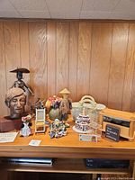 Wide shot showing varied items including bronze bust, metal sculptures, ceramic basket, floral arrangement, and others grouped on a wooden surface against wood panel background.