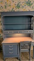 Full view of blue painted Dixie desk with attached bookshelf and chair. Desk has three drawers and a cabinet on the left side. Bookshelf has two open shelves and a light installed below the lower shelf.