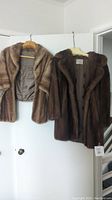 Two brown mink fur coats hanging on hangers against a white wall: one short shawl style and one longer coat.