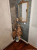 Gold framed mirror mounted on a wall above wooden floor, next to corner with patterned wallpaper and white closet door. Mirror reflects some decor elements and part of the room.
