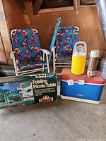 Two folding chairs with colorful patterned fabric, yellow Thermos cooler, blue cooler with red lid, stainless steel and white thermos bottles, and boxed folding picnic table.