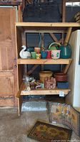 Shelving unit displaying a black overnight luggage bag on top shelf. Middle shelf holds ceramic and plastic pots, watering cans, and decorative swan. Bottom shelf has wicker basket, clay pots, metal spotlights, small human figure sculpture, and black box. Outdoor rugs are in front on the floor.