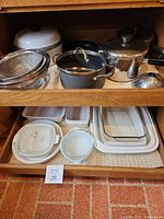 Overview of metal pots, pans, steamers, strainers on top shelf and Pyrex glass and Corning Ware baking dishes in lower shelf.