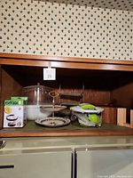 Shelf with tiered serving stand, bagel slicer stand, egg peeler box, clear dome cake cover, and plastic bowl.