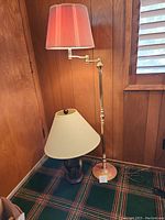 Photo showing both the standing floor lamp with adjustable arm and red pleated lampshade, and the ceramic table lamp with beige shade on green carpet.