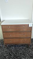 Front view of vintage wooden dresser with white top and three drawers, showing overall condition including wear.