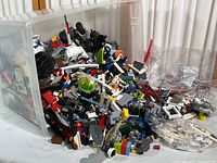 Overspill of mixed LEGO pieces from a clear plastic storage bin showing diverse elements including bricks, plates, wheels, tires, and colors.