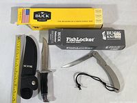 Photo of a fixed blade hunting knife with black handle and nylon sheath next to a fish fillet folding knife with metallic handle and wrist strap, both in their respective Buck Knives boxes.