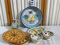 Full set of Italian hand-painted ceramic serving ware including bowl, plates, pitcher, olive dishes, and fish platter shown front-facing on white tablecloth.