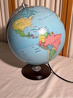 World globe showing Americas with colored countries on a traditional map, mounted on a wooden circular base with metal meridian and electrical cord.