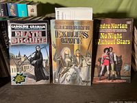 Close-up of three of the visible books on the shelf showing titles and authors: Death Dirge, Exile's Gate, No Night Without Stars.