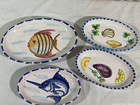 All four oval ceramic dishes displayed on white background showing hand-painted fish and seafood designs with blue and white borders.