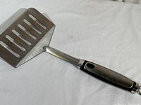 Full view of the outdoor grilling spatula on white background, showing the wide slotted stainless steel blade and black handle.