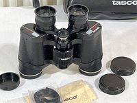 Black Tasco binoculars with two lens caps, cleaning cloth, and strap arranged on white surface