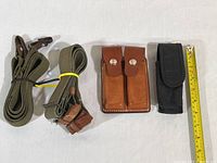 Showing two olive green canvas shoulder straps with leather attachment ends, alongside two pouches: one brown leather double compartment with snap closures, one black canvas with flap closure, measured by adjacent yellow tape measure.