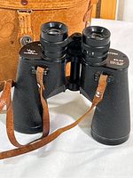 Front view of black textured Saratoga binoculars showing the eyepieces, lenses, and attached leather strap, with the leather carrying case in background.