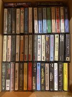 Top view of several rows of audio cassettes in plastic carry case, showing cassette spines with artist names and album titles.