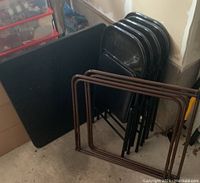 Side view of the folded black table, stacked folding black chairs and two brown metal folding tabletop supports