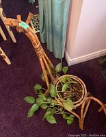 Front and side views of a giraffe-shaped plant holder made of cane/rattan holding a potted vine plant with green leaves on dark purple carpet.