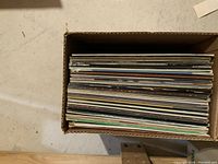 Top view of about 40 assorted LP vinyl records stacked side by side in a cardboard box.