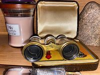 Front view of opera glasses with brass and mother of pearl body, resting on vintage playing cards, next to a candle and rock.