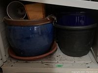 Photo showing large blue glazed ceramic pot with terracotta saucer and black plastic pot on shelf