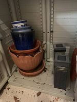 Photo of four planting pots stacked, showcasing a large dark blue glazed pot, a smaller blue and white decorative pot, two terracotta pots, and two rectangular standing garden lights next to them.