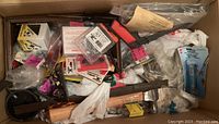 Top view of cardboard box containing an assortment of tools and hardware fasteners including claw hammer, screws and nails in packaging with pink auction tags visible.