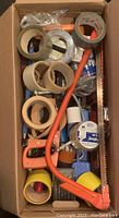 Box with multiple rolls of tape, an orange hand saw, and other small tools organized on top in a cardboard box.