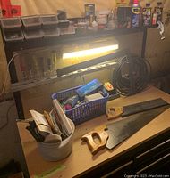 View of entire work surface showing two hand saws with wooden handles, blue basket with assorted tools, white container with tools and parts, coiled black cable or hose, mounted light, pegboard with screwdrivers and other tools, and several spray cans including WD-40.