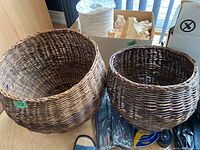 Photo of two round natural brown wicker baskets, one larger and one smaller, placed on wooden floor with other household items partially visible.
