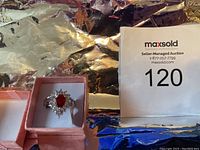 Front view of sterling silver ring with a prominent red gemstone surrounded by smaller clear stones in pink box.