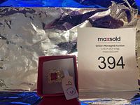 Ring displayed in a red jewelry box showing the size tag '9'. Central red stone with surrounding clear stones and detailed gold tone design.