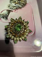 Close-up of vintage brooch showing central green stone, amber stones, and textured gold-tone metal petals with green stone tips.